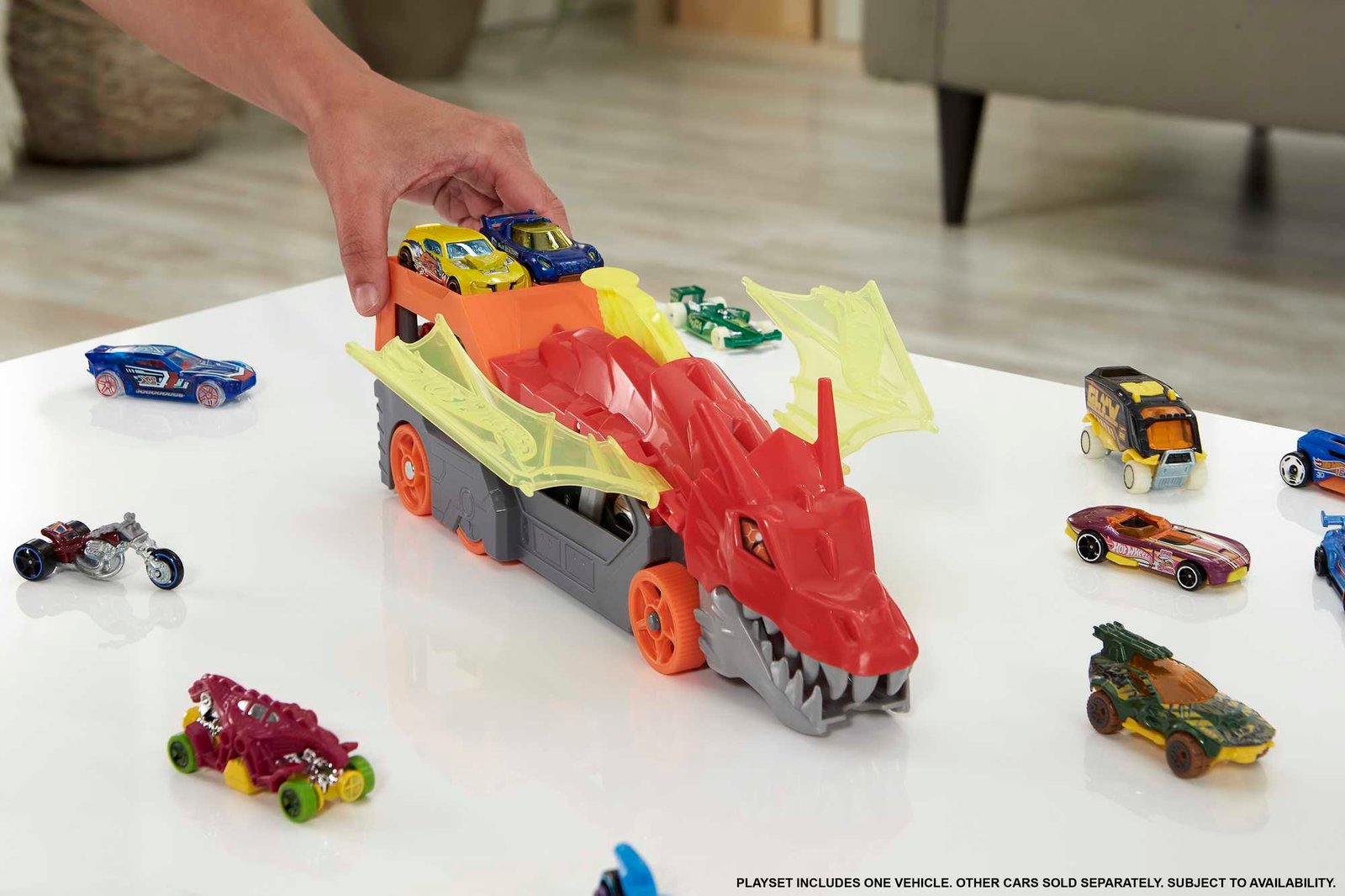 Ranka deda mašinėles ant drakono transporterio, aplink išdėlioti Hot Wheels automodeliukai