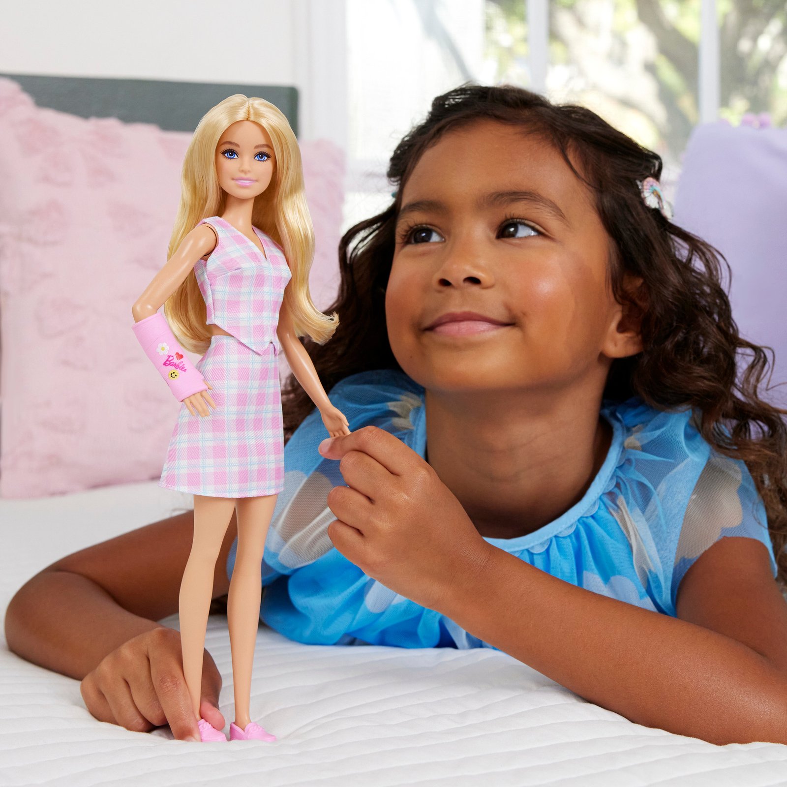 Mergaitė ant lovos žaidžia su Barbie lėle, vilkinčia languotą aprangą