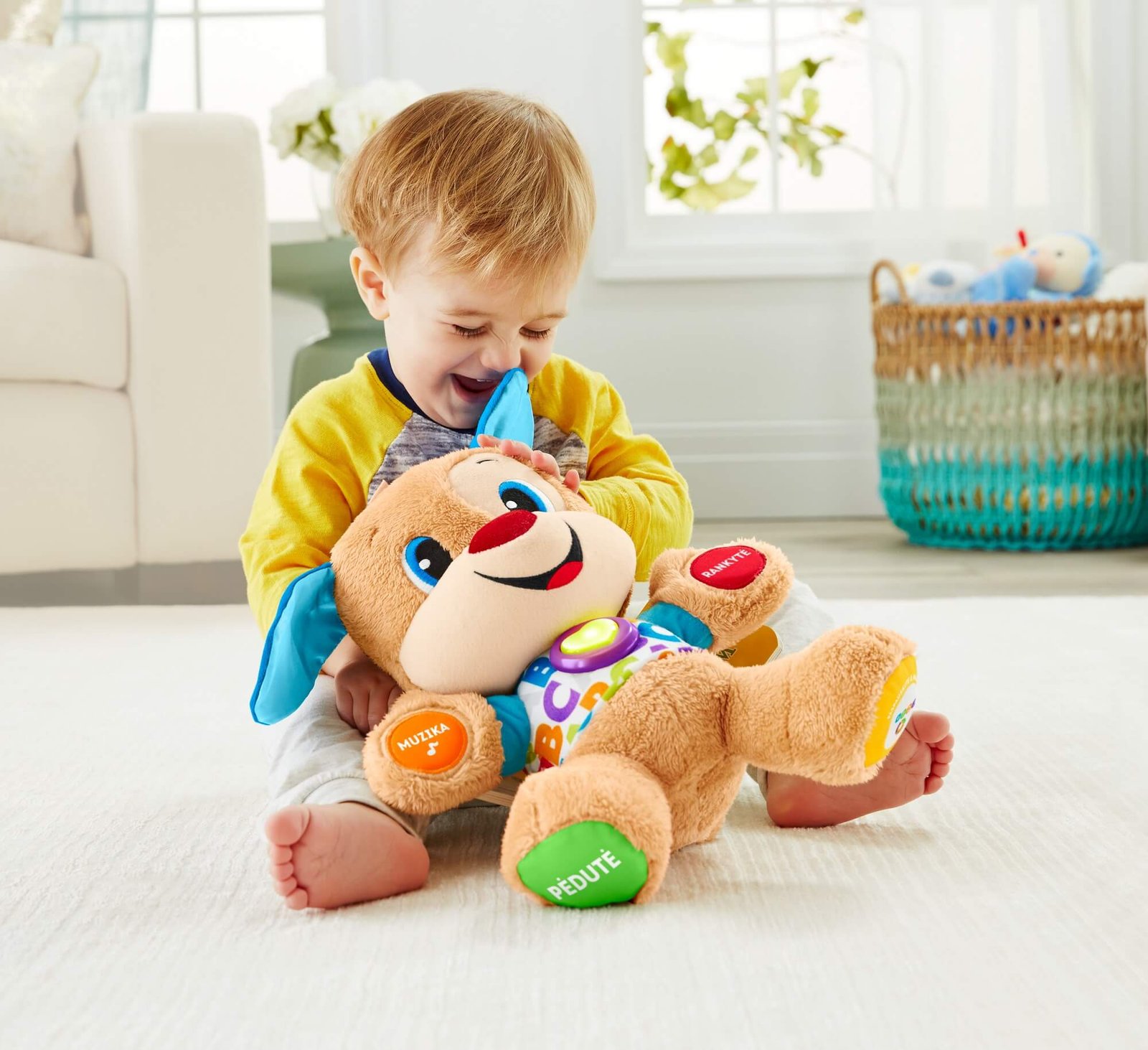 Mažylis sėdi ant kilimo ir apkabina Fisher-Price mokomąjį šuniuką